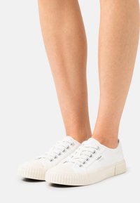 White canvas sneakers with lace-up design, rubber toe cap, and textured sole. Features silver eyelets and brand logo on outer side.