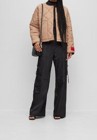 Kvinde iført tanfarvet quiltet jakke, sorte cargobukser, sorte sandaler og en sort skuldertaske mod en ensfarvet grå baggrund.