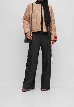 Femme portant une veste matelassée beige, un pantalon cargo noir, des sandales noires et un sac à bandoulière noir sur un fond gris uni.