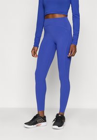 Atletske tajice u kraljevsko plavoj boji s visokim strukom, bez šavova i glatkom teksturom, u kombinaciji s crnim sportskim cipelicama.