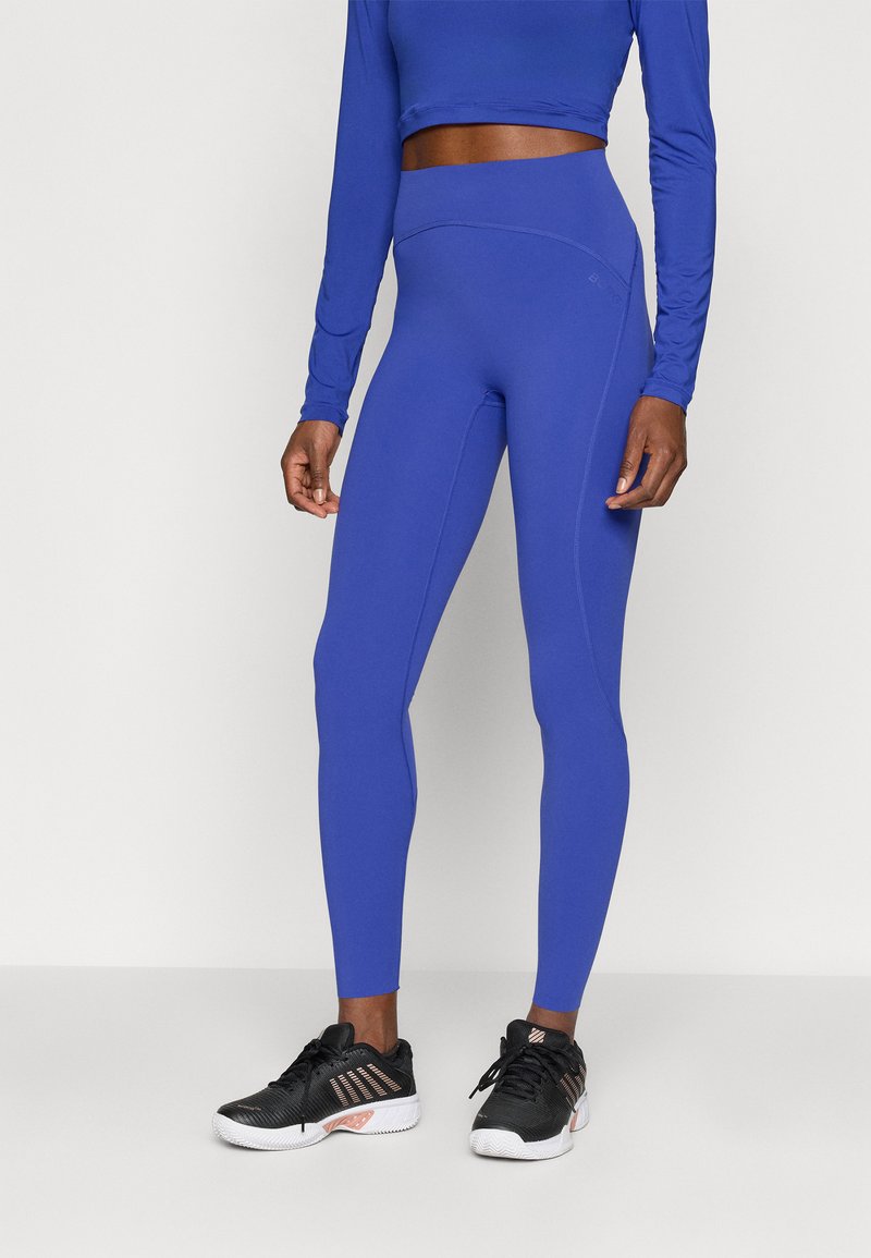 Atletske tajice u kraljevsko plavoj boji s visokim strukom, bez šavova i glatkom teksturom, u kombinaciji s crnim sportskim cipelicama.