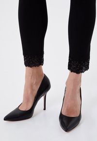 Zapatos de tacón alto de cuero negro con un diseño de punta afilada, delante de unos jeans negros hasta el tobillo con detalles de encaje y lentejuelas en el dobladillo.