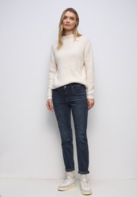Kremowy, puszysty sweter z wysokim kołnierzem, zestawiony z ciemnoniebieskimi, dopasowanymi dżinsami i białymi trampkami z szarymi akcentami. Prosty fason z podwiniętymi mankietami.