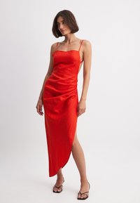 Abito rosso senza maniche con spalline sottili, orlo asimmetrico e texture arricciata, realizzato in un tessuto leggero. Indossato con scarpe nere con cinturini.