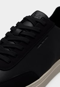 Baskets en cuir noir avec lacets noirs, logo embossé sur la languette et le côté, et une semelle en caoutchouc beige clair.