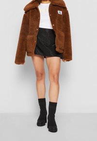 Bruine faux fur jas met een kraag, wit topje, zwarte satijnen rok en zwarte kniehoge laarzen met hak.