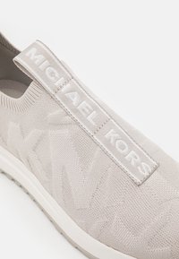 Chaussure à enfiler gris clair en matériau tricot texturé, dotée d'un large onglet logo blanc portant "MICHAEL KORS" sur le côté et d'une semelle blanche.