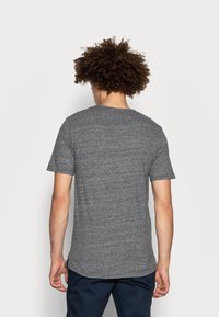 Mand med krøllet hår iført en ensfarvet grå t-shirt med korte ærmer og mørke bukser, vist fra ryggen mod en ensfarvet hvid baggrund.