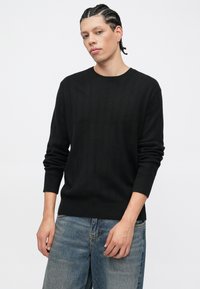 Μαύρο πλεκτό πουλόβερ με ριγέ υφή, μακριά μανίκια, στρογγυλή λαιμόκοψη και μανσέτες, συνδυασμένο με ανοιχτό μπλε τζιν παντελόνι.