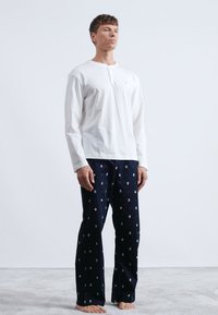 Chemise henley blanche à manches longues avec un col rond, associée à un pantalon de pyjama bleu marine orné d'un petit motif circulaire.