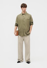 Man wearing olive green button-up shirt and beige wide-leg pants, holding a small brown leather case, standing against plain background.