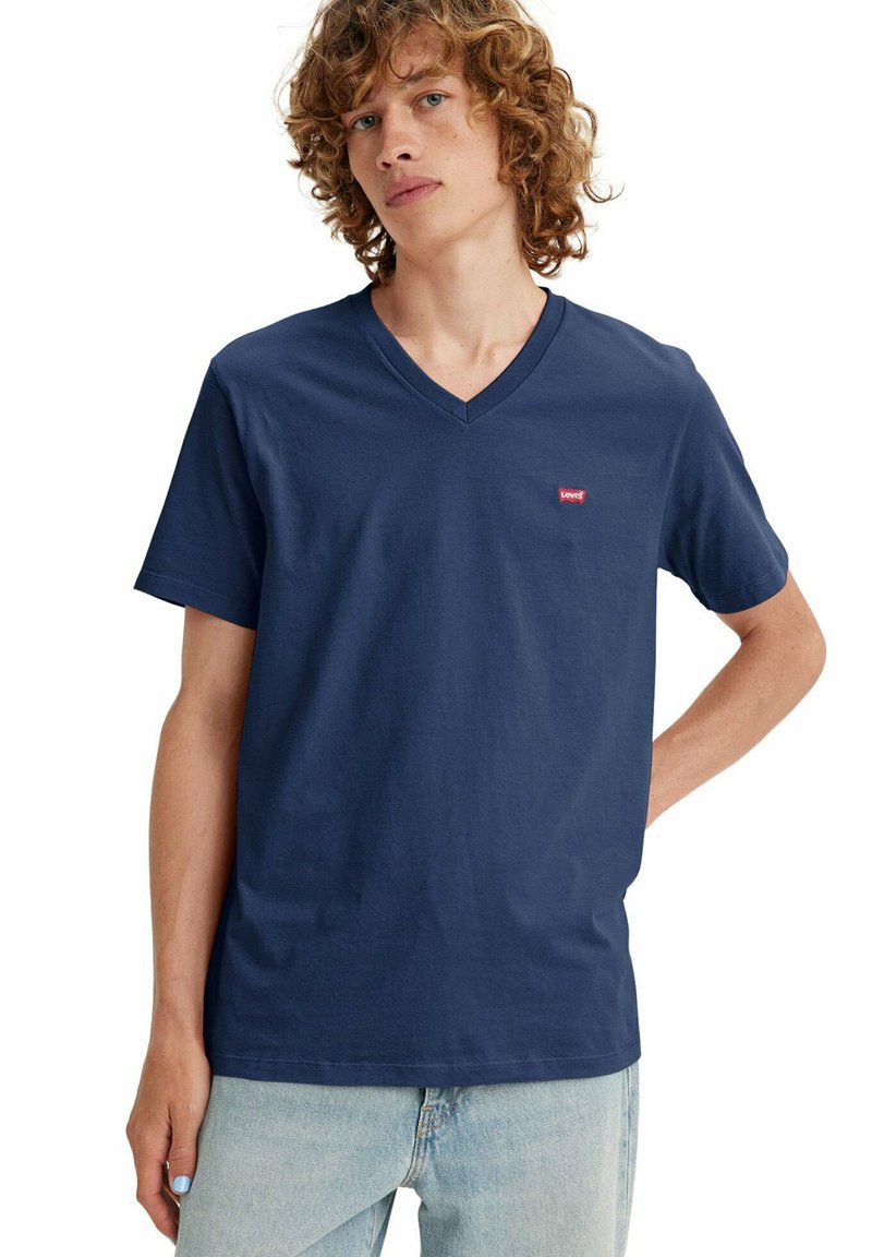 Junger Mann mit lockigem Haar, der ein dunkelblaues T-Shirt mit V-Ausschnitt und hellblaue Jeans trägt, steht mit einer Hand in der Hüfte.