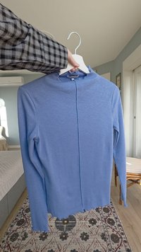 Haut bleu clair à manches longues avec une texture côtelée, ouverture à l'avant, encolure arrondie et ourlet brut en bas. Suspendu à un cintre blanc.