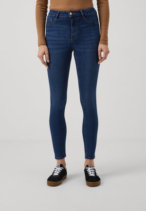 Vrouw die donkere blauwe skinny jeans, een bruin shirt met lange mouwen en zwarte sneakers met witte veters draagt, staand tegen een neutrale achtergrond.