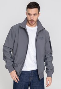 Graue Softshell-Jacke mit hohem Kragen, durchgehendem Reißverschluss und seitlichen Taschen. Mit einem Logo auf der Brust und einem strukturierten Innenfutter.