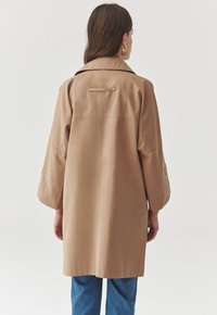 Trench-coat beige avec une coupe décontractée, mettant en avant un détail de dos et des manches larges. Porté avec un pantalon en denim bleu. Texture de tissu lisse.