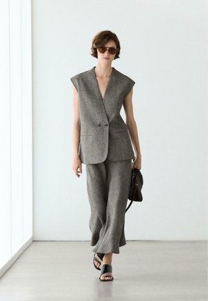 Donna che indossa un blazer grigio senza maniche e pantaloni ampi abbinati, con una borsa nera, che cammina in una stanza bianca minimalista.