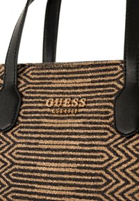 Borsa a tracolla in marrone e beige a motivo con manici in pelle nera. Presenta un logo dorato "GUESS" e un tessuto testurizzato con linee geometriche.