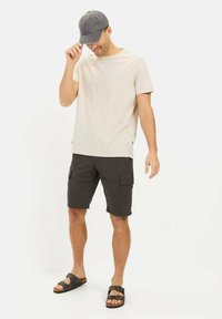 Beige kortærmet skjorte, mørke cargo shorts og grå kasket. Sandaler med brune stropper. Model der står med hånden på kasketten og smiler.