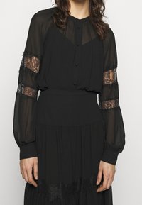 Schwarzes Langarmkleid mit Spitzenakzenten, Knopfleiste vorne, geraffter Taillenteil und strukturiertem Stoff, mit transparenten Ärmeln und einem fließenden Rock.