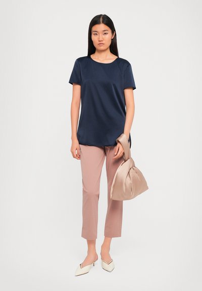 Woman wearing navy short-sleeve blouse, dusty pink cropped pants, white heels, holding a large beige soft handbag by her side.