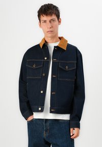 Donkerblauwe denim jacket met een corduroy kraag, twee borstzakken, knopen aan de voorkant en stikseldetails. Draag het over een wit shirt.