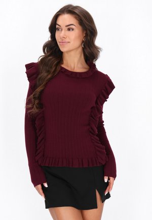 Frau mit langen, welligen Haaren, die einen bordeauxroten Rüschenpullover und einen schwarzen Rock mit vorderem Schlitz trägt, schaut seitlich.