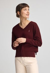 Bordeaux v-ringad tröja i mjukt material, med en figursydd nederkant och en liten broderad logotyp på bröstet. Bärs med ljusa byxor.