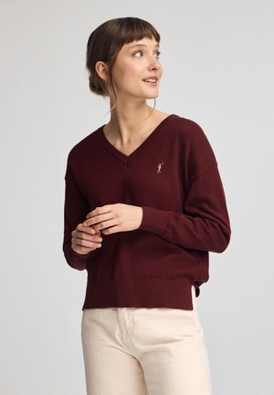 Bordeaux v-hals sweater in zachte stof, met een aansluitende onderzoom en een klein geborduurd logo op de borst. Draagt met lichtgekleurde broek.