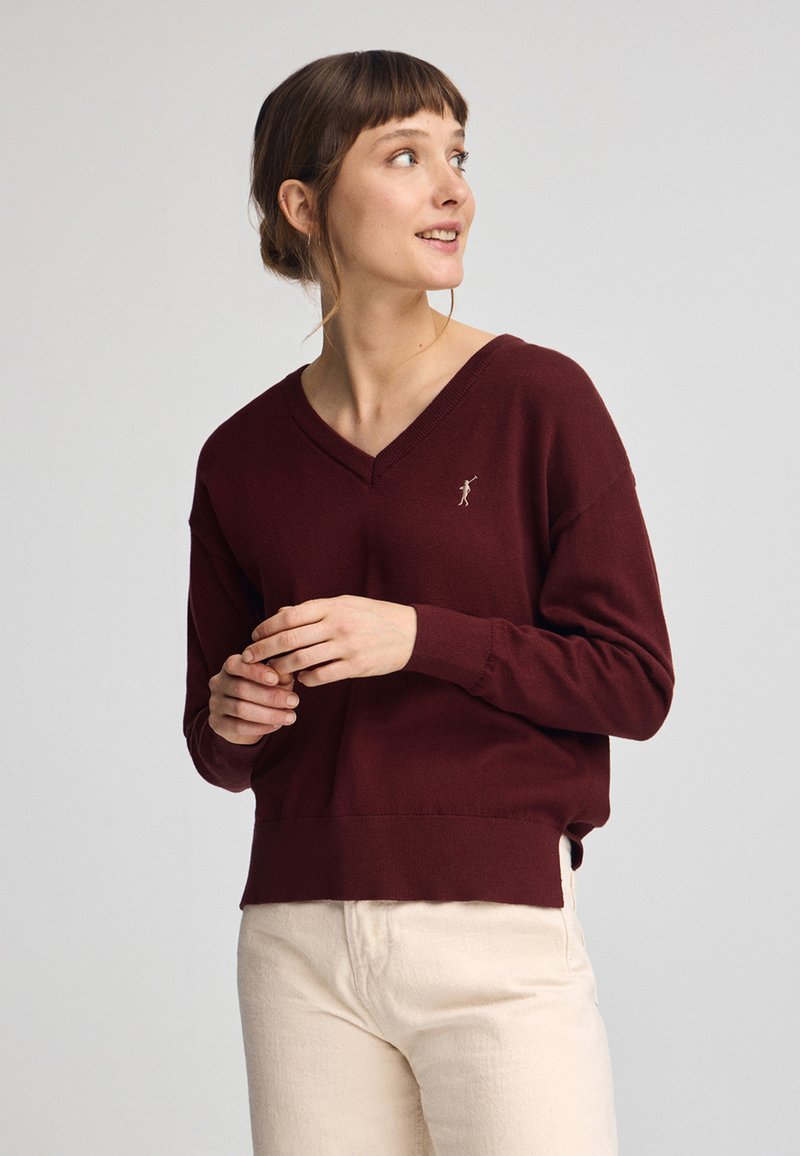 Bordeaux v-ringad tröja i mjukt material, med en figursydd nederkant och en liten broderad logotyp på bröstet. Bärs med ljusa byxor.