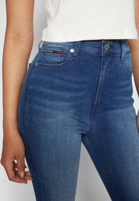Jeans de mezclilla azul de tiro alto con una textura suave, un leve desgaste y cierre de botón metálico, con un toque de acento rojo en el bolsillo.