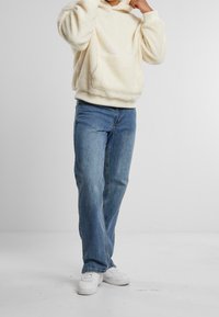 Hoodie di colore crema, morbido e peloso, con tasca frontale, abbinato a jeans dritti azzurri e sneakers bianche su uno sfondo neutro.