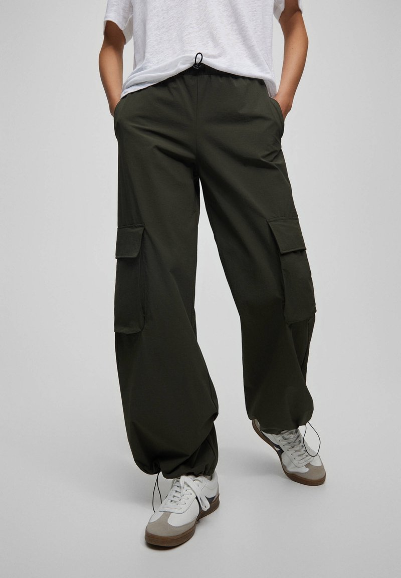 Personne portant un pantalon cargo vert foncé avec des poignets élastiques, des baskets blanches et un haut blanc à manches courtes, les mains dans les poches, debout sur un sol gris.