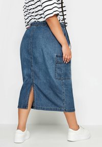 La falda midi de mezclilla presenta una cinturilla elástica, un bolsillo de parche, aberturas laterales y un color azul desgastado. Se combina con zapatillas blancas.