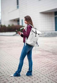 Světlě šedý batoh s rolovacím víkem a nastavitelnými popruhy. Model má na sobě vínovou bundu, modré zvonové džíny a bílé tenisky, drží smartphone.