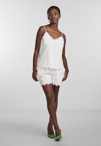 Hvit brodert topp med lagdelt design og bølgete kanter, kombinert med matchende shorts. Modellen har på seg grønne hæler.