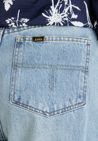 I jeans in denim azzurro chiaro presentano una tasca posteriore con un dettaglio di cucitura gialla e un'etichetta del marchio Lois nera con testo giallo.