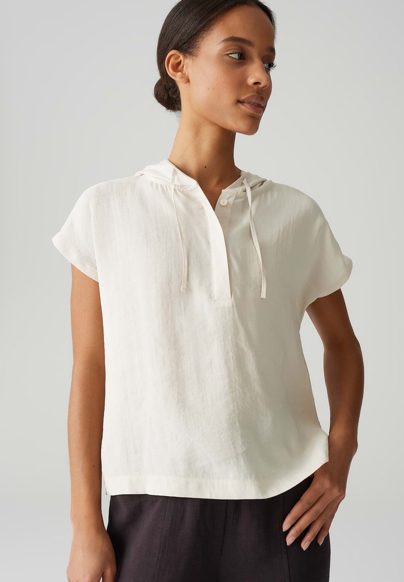 Blusa corta a maniche corte color beige chiaro con cappuccio, patta e coulisse. Realizzata in tessuto testurizzato, presenta una vestibilità rilassata e orlo dritto.
