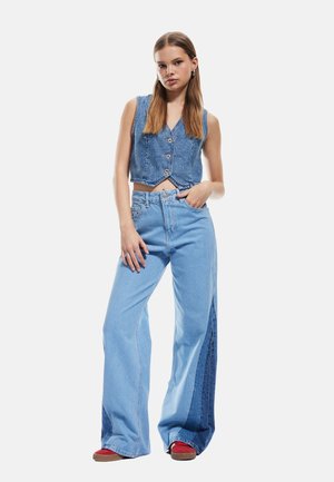 Denim crop top met een knopen voorkant en een strik in de taille, gecombineerd met wijde jeans met een donkerdere denim zijpaneel. Rode schoenen.