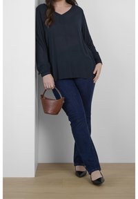 Blouse à manches longues bleu foncé avec un col en V, portée avec un jean en denim foncé. Tient un petit sac à main en cuir marron avec une poignée incurvée.