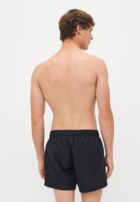 Shorts de bain noirs avec une taille élastique et une poche arrière. Texture lisse et design arrivant au genou, vus de dos.