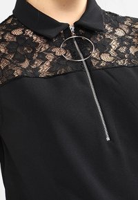 Haut noir avec un panneau en dentelle à motif floral, un col et une fermeture éclair avec un accent en anneau circulaire. Tissu lisse en dessous de la dentelle.