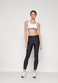 Balta sportinė liemenėlė su plačia juoda Under Armour juosta, derinama su juodomis legging'ais, turinčiais glotnų tekstūrą ir prigludusį dizainą.