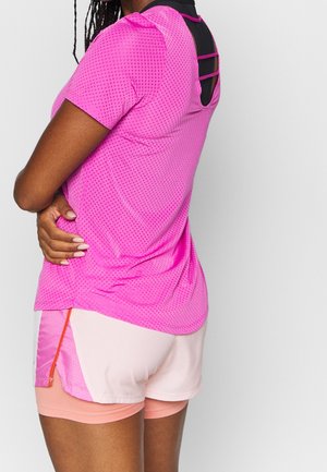 Femme portant un haut court à manches courtes perforé rose vif et un short superposé rose clair, debout, les bras croisés, devant un fond uni.