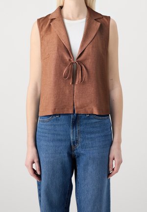 Sleeveless brown cropped vest with lapels tied at the front, worn over a white top and paired with blue high-waisted denim jeans.