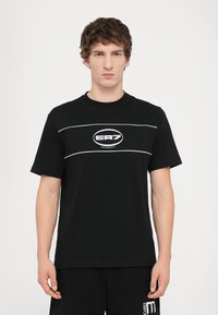 Černé bavlněné tričko s krátkým rukávem, na kterém je uprostřed bílé logo s rámečkem a dvě horizontální bílé čáry přes hrudník.