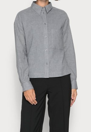 Kobieta w szarej koszuli na guziki z kołnierzykiem i przednią kieszenią na piersi, w zestawieniu z czarnymi spodniami na jednolitym tle.