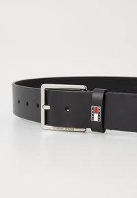 Zwarte synthetische riem met een rechthoekige zilveren gesp. Heeft gaatjes voor aanpassing en een logo-accent nabij de gesp. Gladde textuur.
