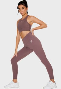 Reggiseno sportivo mauve e leggings a vita alta realizzati in tessuto elastico. Accentuati con un logo in argento. Modella in posa con una gamba in avanti.