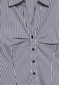 Chemise vichy bleu et blanc avec col large, détail froncé à la poitrine et quatre boutons bleu foncé au centre devant.